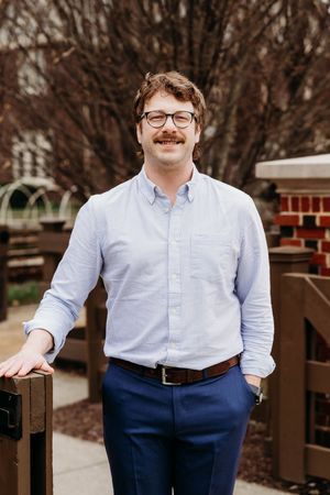 Man with mustache and glasses smiling outdoors, wearing blue shirt and navy pants, leaning on wooden fence.