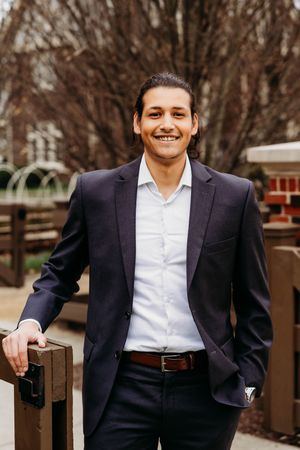 Smiling young man in navy blazer and white shirt posing outdoors near wooden railing with bare trees in background.