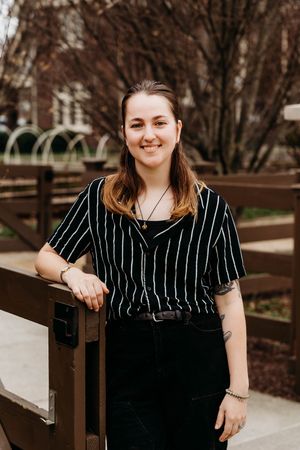 Young woman smiling in black striped top and dark jeans, leaning on wooden fence outdoors in spring.