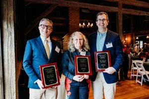 Three professionals smile holding award plaques at an indoor ceremony venue with warm lighting.