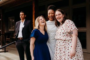 Three women laughing and embracing outdoors at a formal event, with a man in a suit standing in the background.