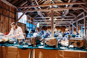 A woman serves food from chafing dishes at a buffet inside a rustic wooden barn venue during a catered event.