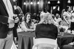 Guests mingle and converse at round tables during an indoor event in a rustic venue, captured in black and white.