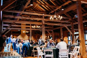 Guests dine at round tables inside a rustic barn wedding reception venue with exposed wooden beams and chandeliers.