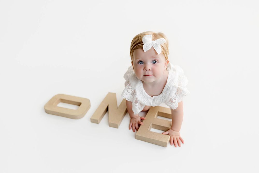 Baby girl in white dress poses with wooden ONE letters for her first birthday milestone photo session.