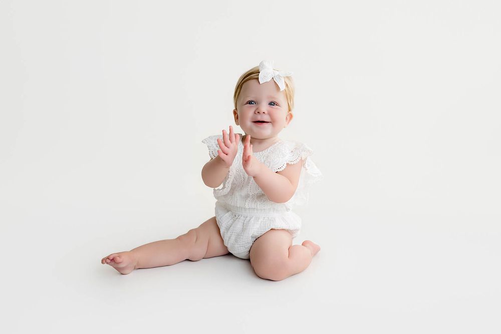 Smiling baby girl in white lace romper and bow headband clapping hands on white background.