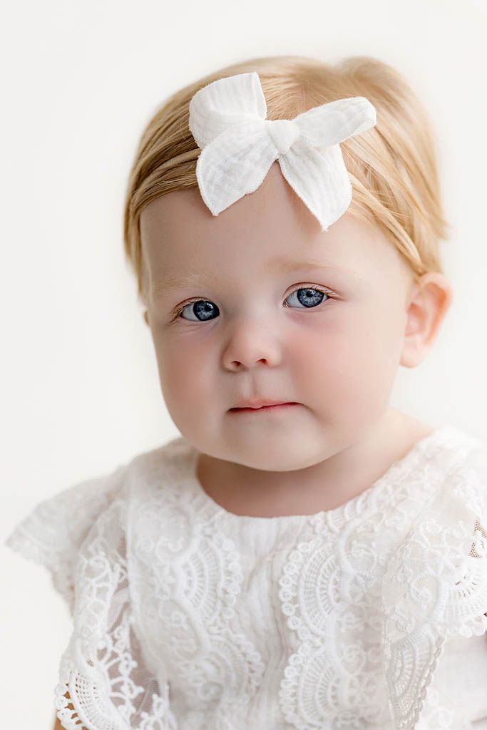 Adorable baby girl with blue eyes wearing a white lace dress and white bow headband against a light background.