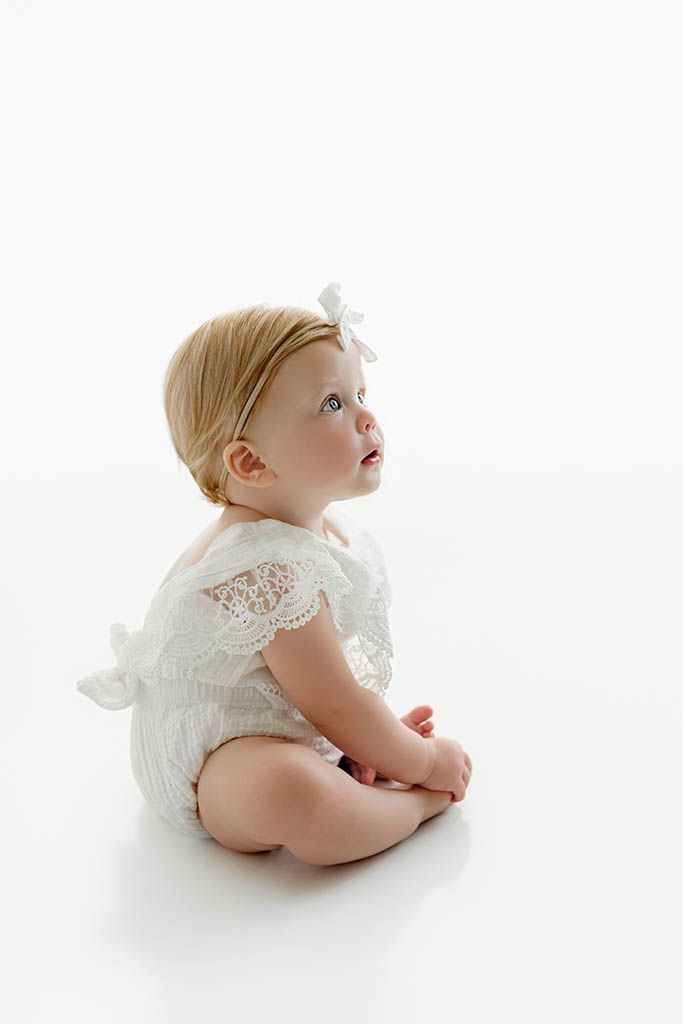 Adorable baby girl in white lace romper and bow headband sitting on white background looking up.
