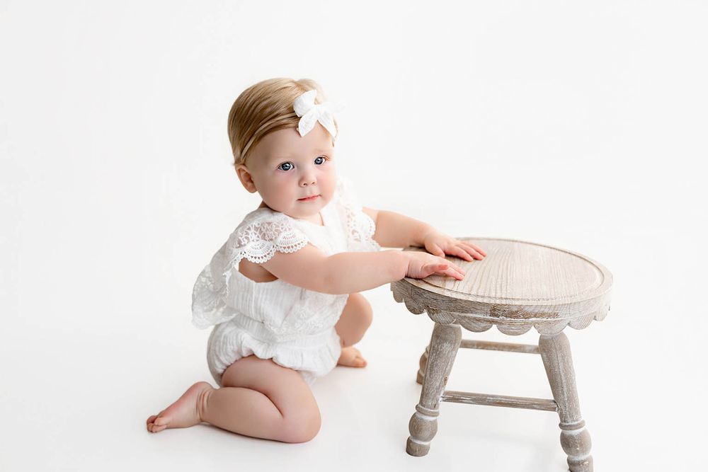 Adorable baby girl in white lace romper with bow headband kneeling beside a small wooden stool.