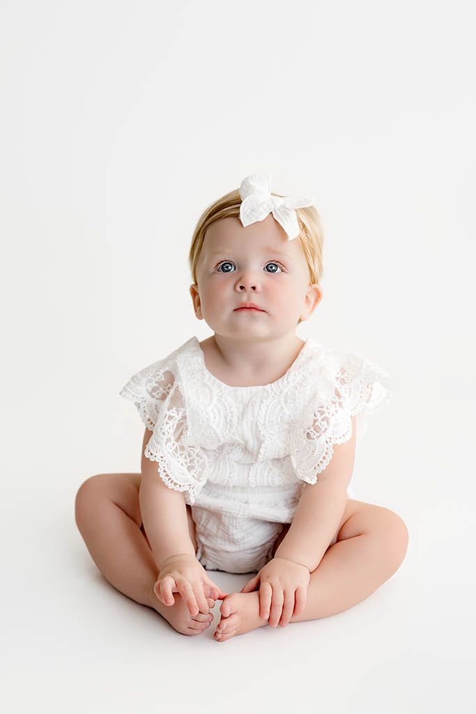 Adorable baby girl in white lace romper and bow headband sitting cross-legged on white background.