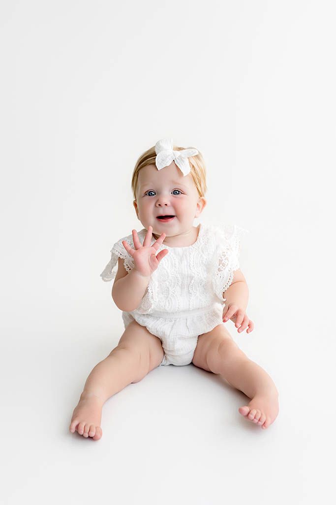 Smiling baby girl in white lace romper and bow headband sitting on white background.