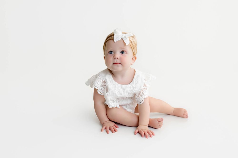 A cute baby girl in a white lace romper and bow headband sits on a white background, looking up curiously.
