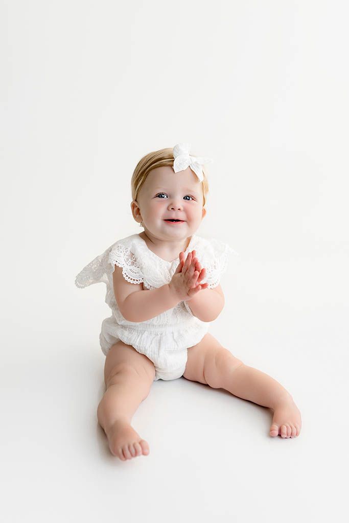 Happy baby girl clapping hands in white lace romper and bow headband on white background.