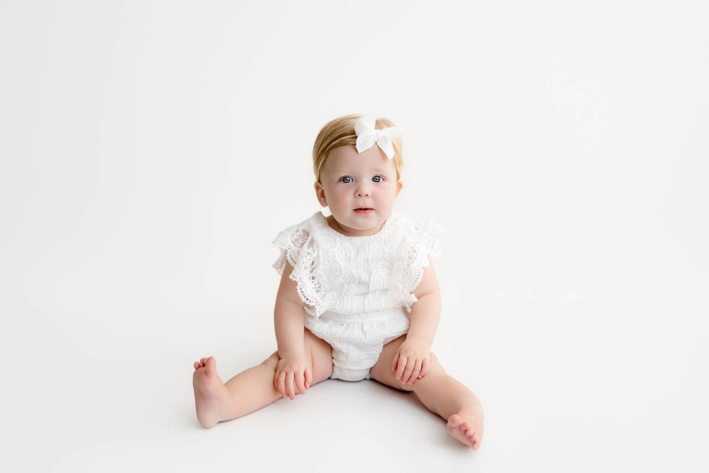 Cute baby girl sitting on white background wearing a white lace romper and bow headband.
