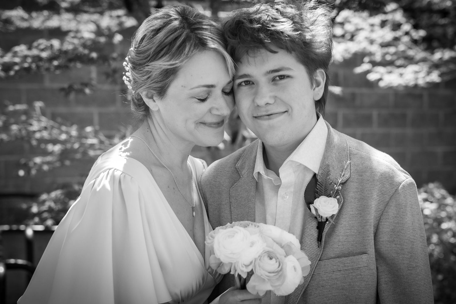Black and white wedding photo of smiling bride and groom holding bouquet outdoors near brick wall.