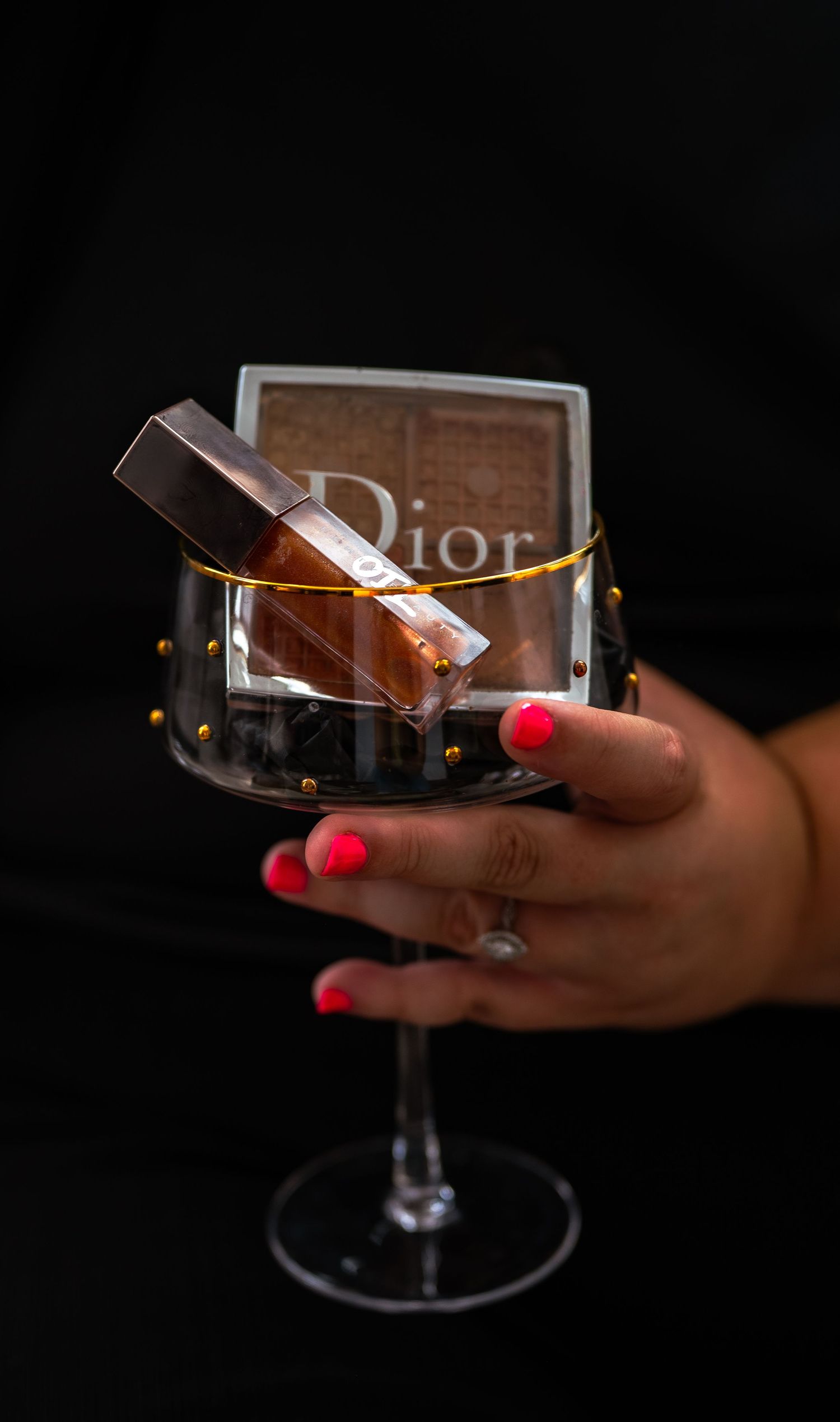 A hand with red nails holds a crystal wine glass containing Dior makeup products against a dark background.