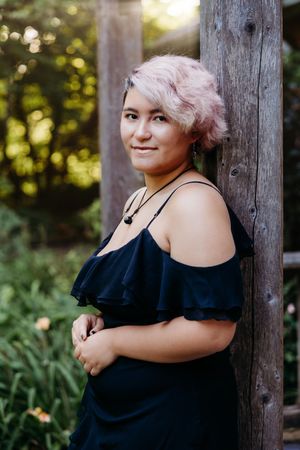 Young woman with short pink hair in a black off-shoulder dress leaning against a wooden post outdoors.