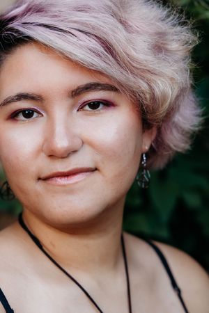 Young woman with short pastel pink hair, subtle makeup, and dangling earrings smiling softly outdoors.