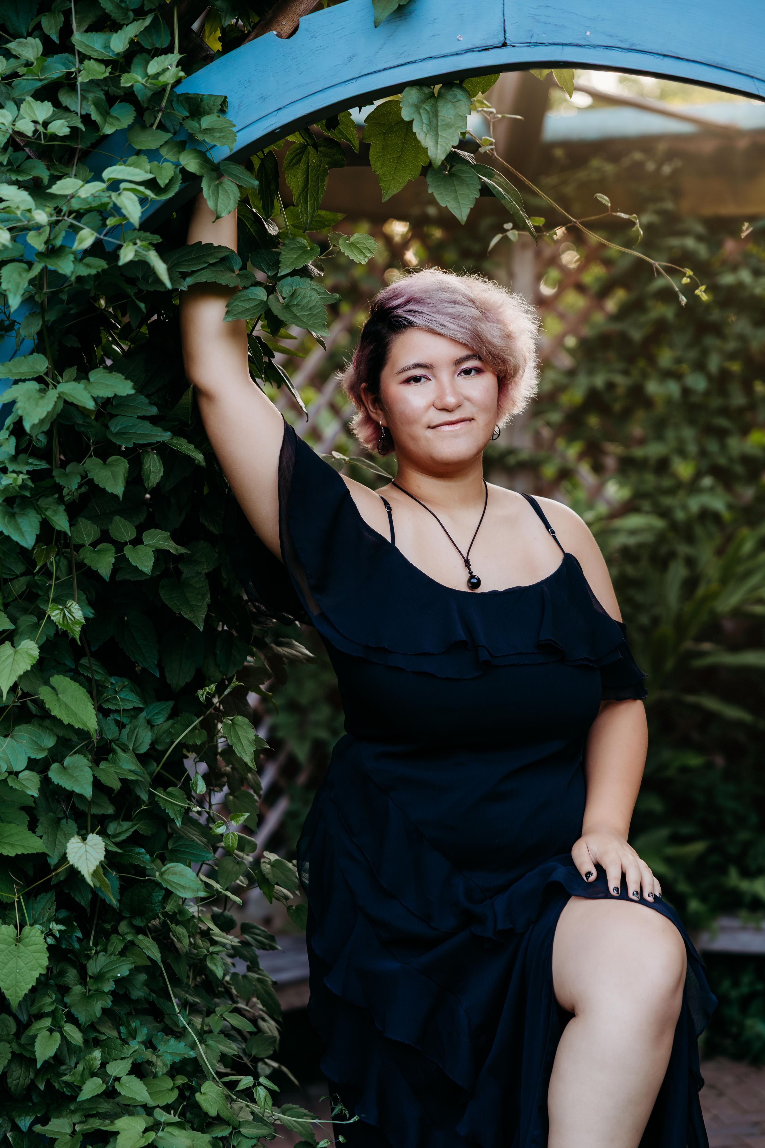 A plus-size woman in a black dress poses gracefully beside a blue arch covered in lush green ivy outdoors.
