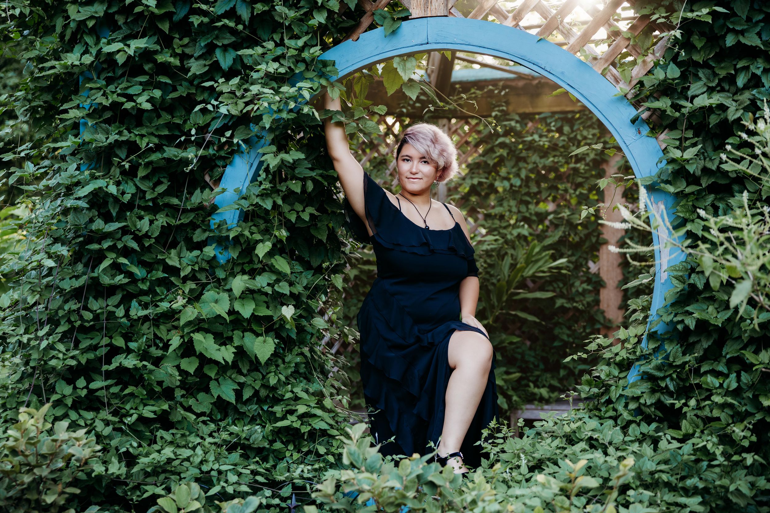 Woman in black dress posing in circular garden archway surrounded by lush green ivy foliage.