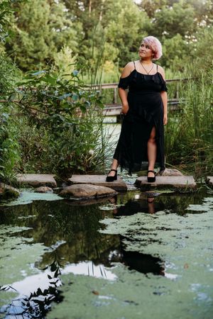 Woman in black dress standing on stepping stones over a serene pond surrounded by lush green garden foliage.