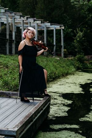 Woman in black dress playing violin on wooden dock beside green algae-covered pond surrounded by lush forest.