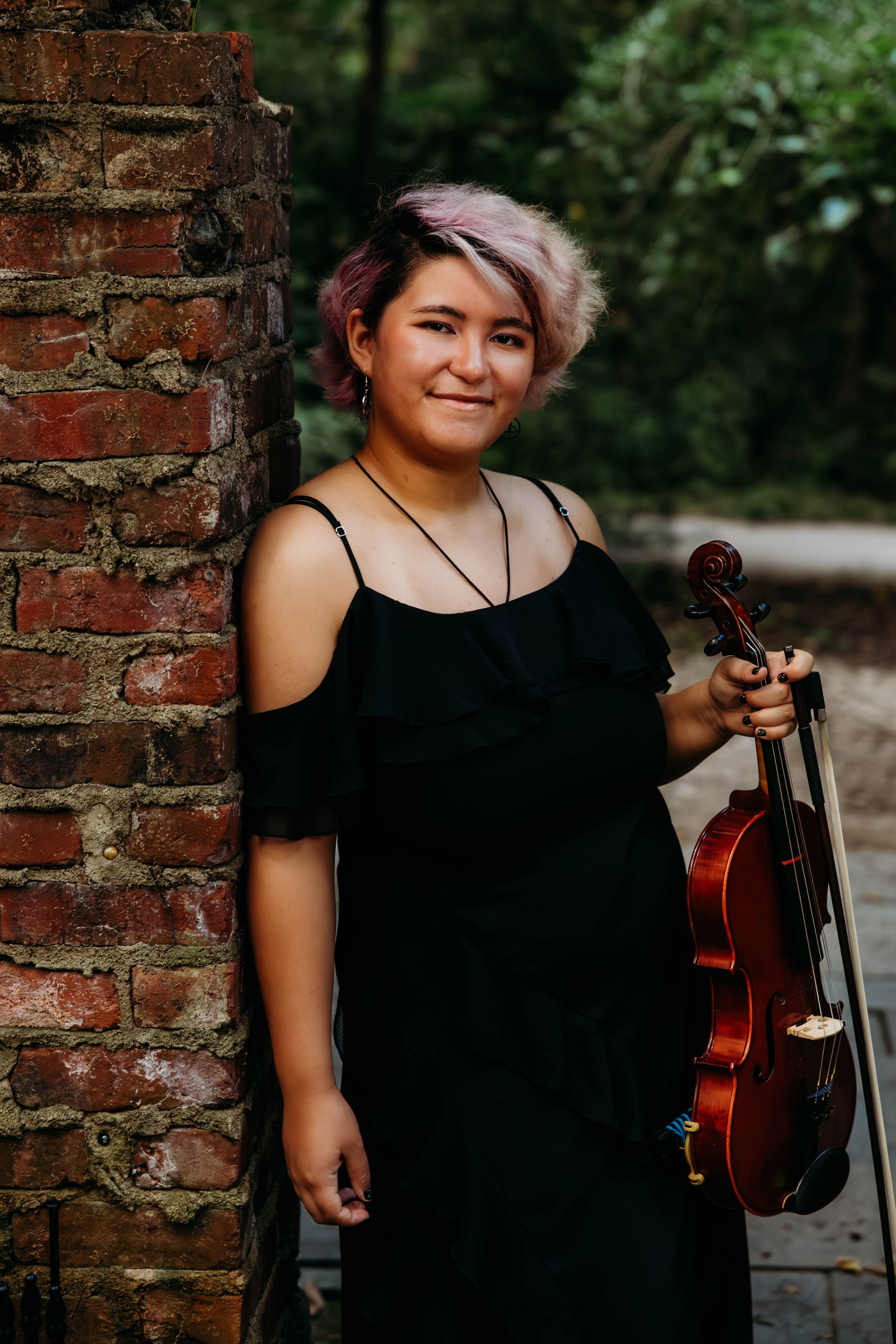 Young woman in black dress holding a violin, smiling beside a rustic brick wall outdoors.