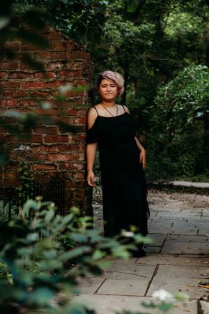 A young woman with pink hair in a black off-shoulder maxi dress poses by a rustic brick wall surrounded by lush greenery.