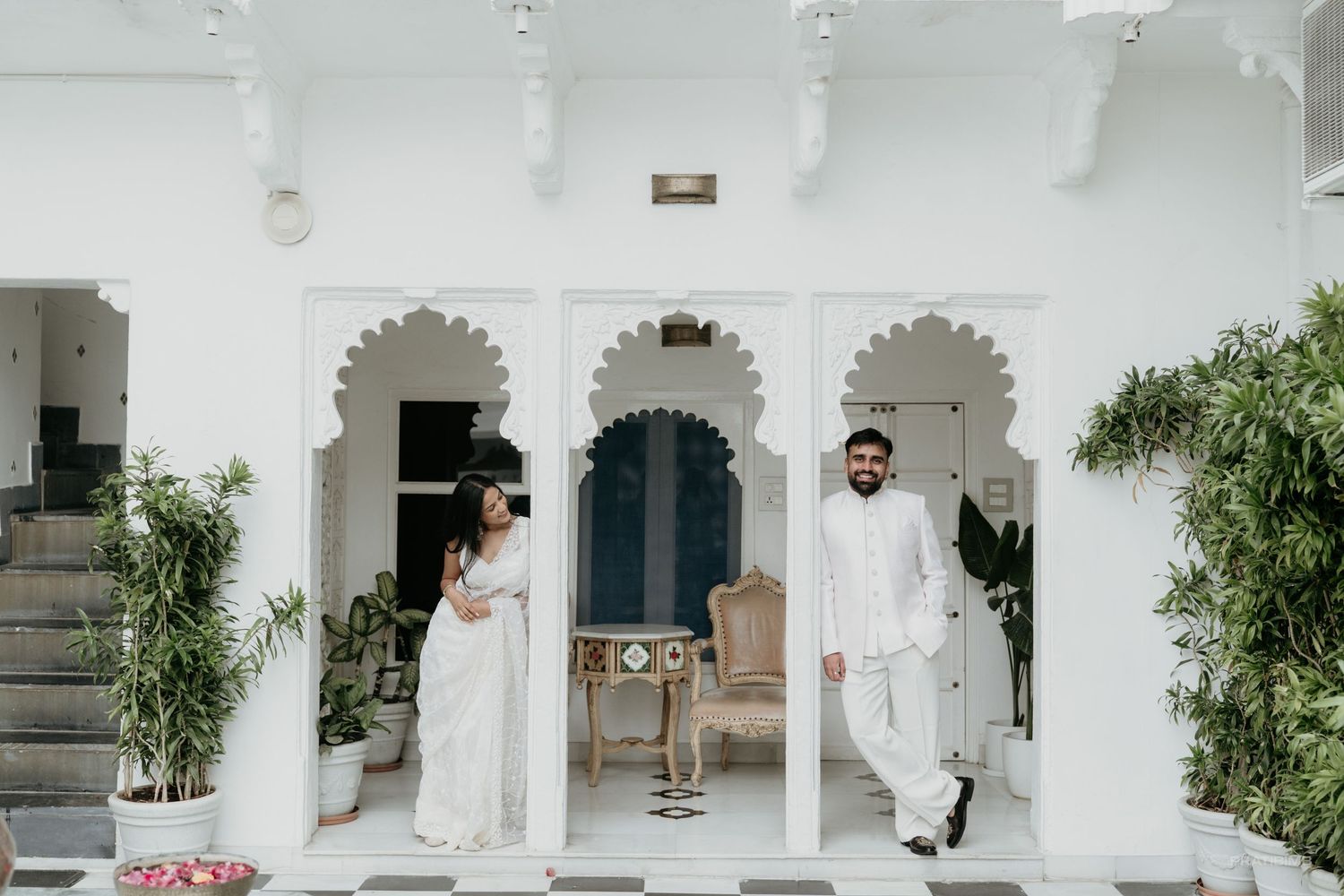 Couple in white traditional attire posing in elegant white arched venue during Indian wedding ceremony.
