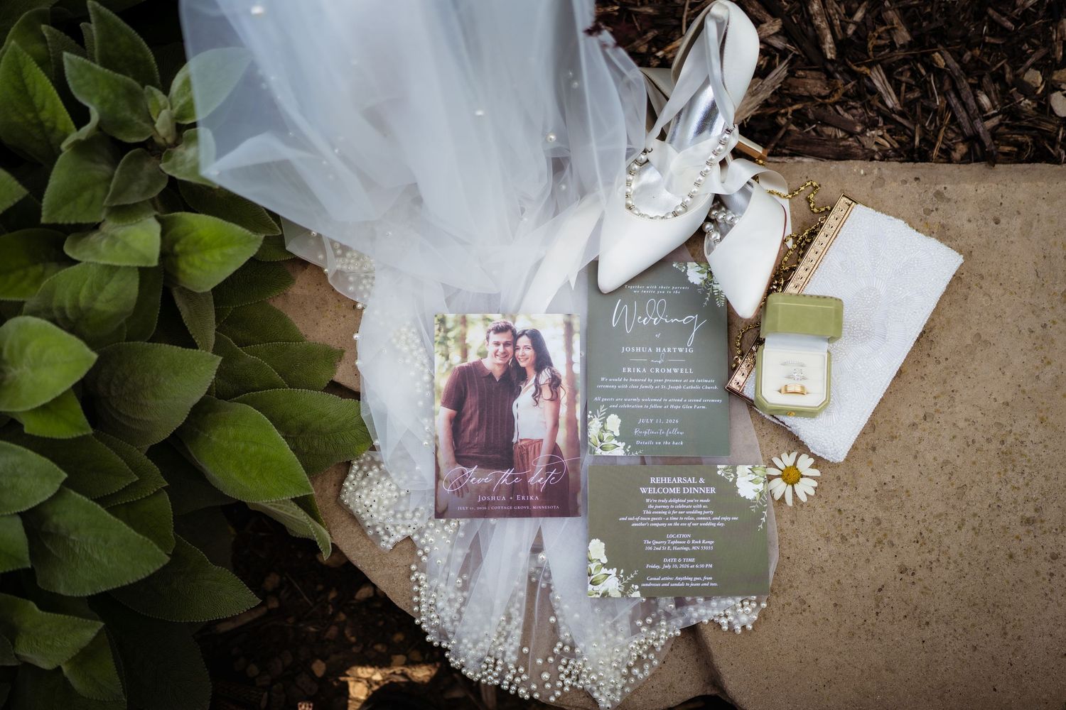 Wedding flat lay with invitation, photo card, white heels, veil, ring box, and daisies on stone surface.