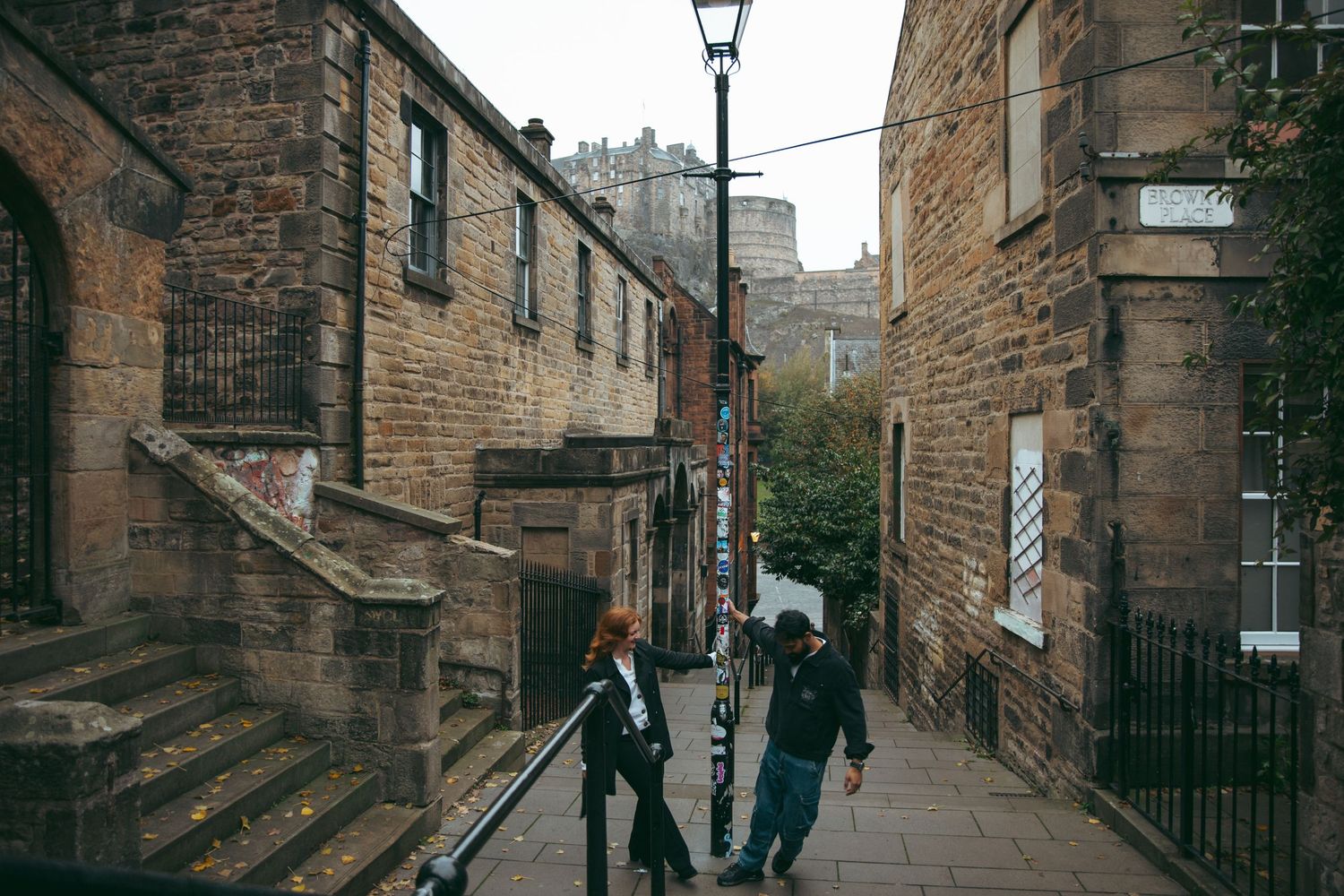 Couple walking through a historic cobblestone alley in Edinburgh with the castle visible in the background.