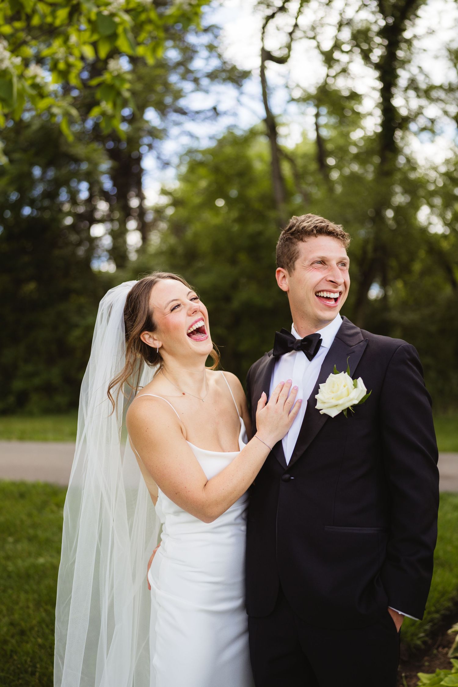 Joyful bride and groom laughing outdoors, bride in white gown with veil, groom in black tuxedo with white boutonniere.
