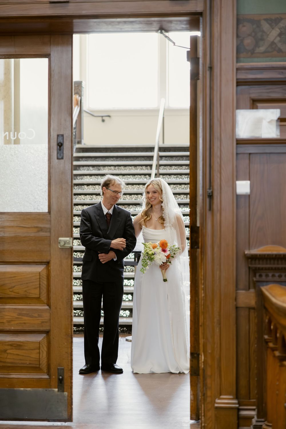 Bride in white gown and veil holding bouquet stands with escort at courthouse entrance before ceremony.