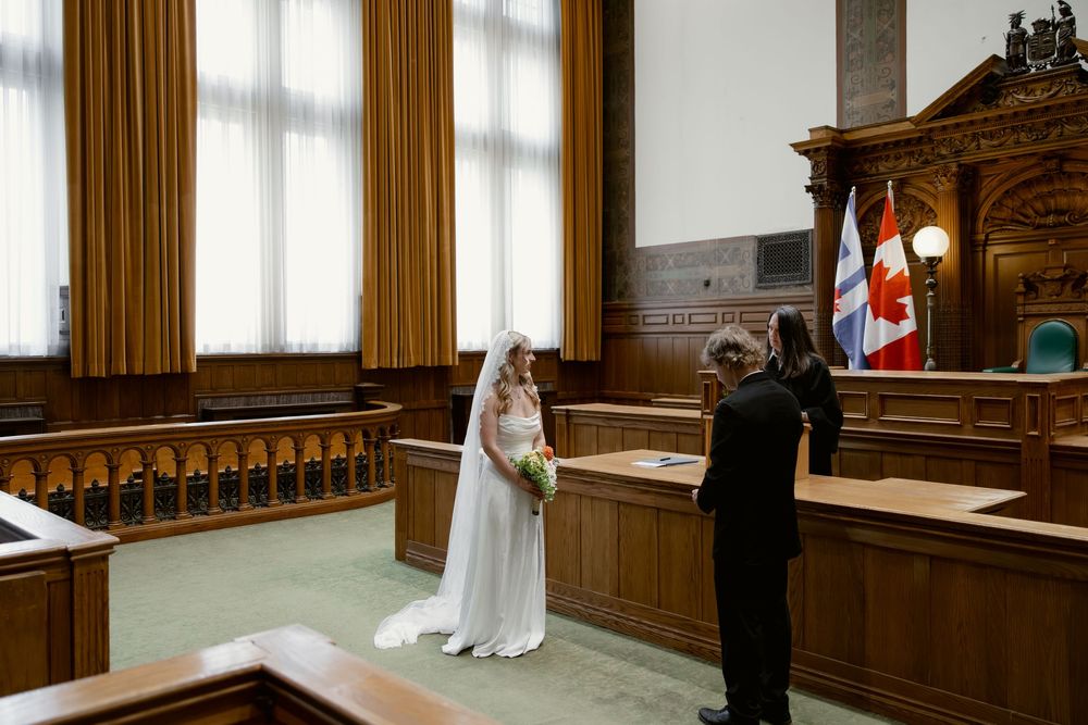 Bride in white gown and veil stands before officiant in a wood-paneled courtroom ceremony.