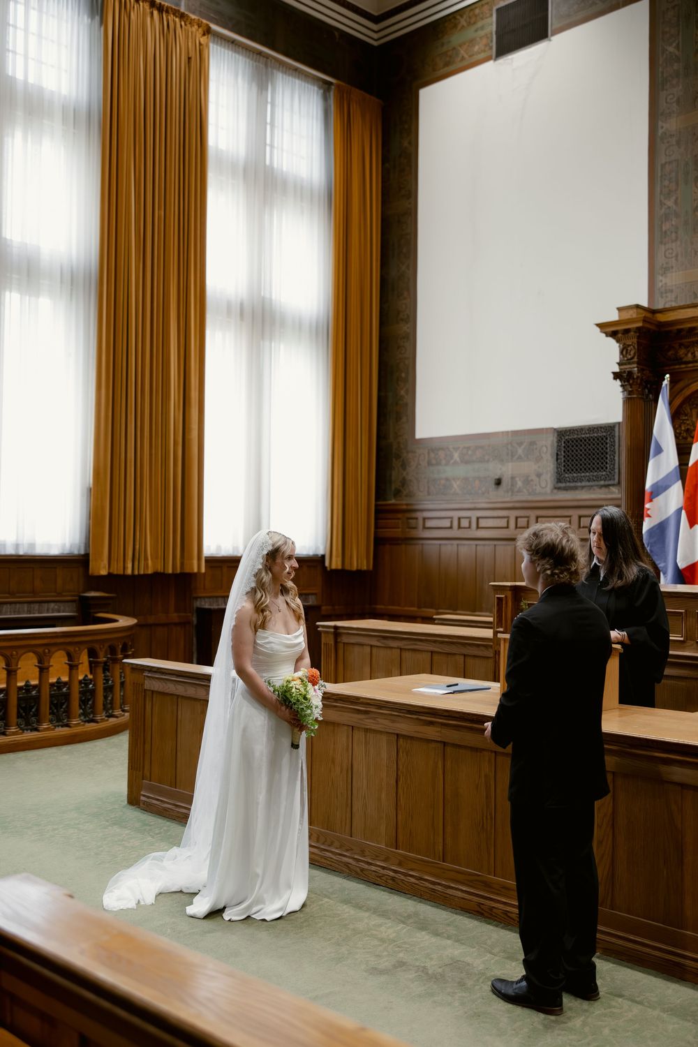 Bride in flowing white gown holds bouquet while listening to officiant during courthouse wedding ceremony.