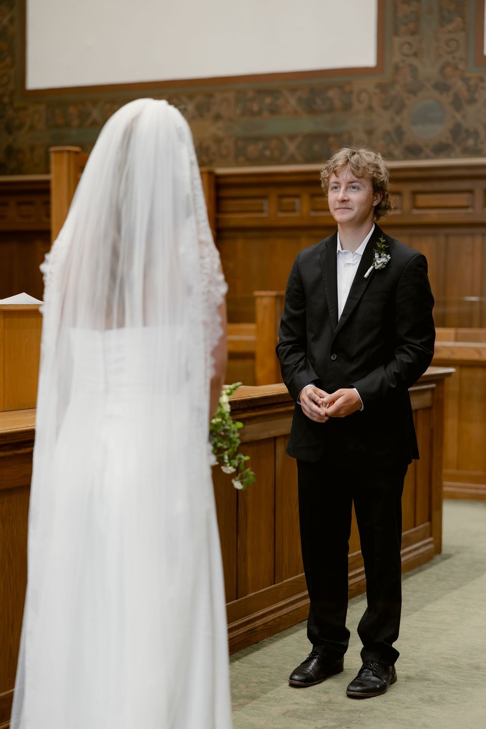 Groom in black suit gazes at veiled bride during intimate courthouse wedding ceremony.