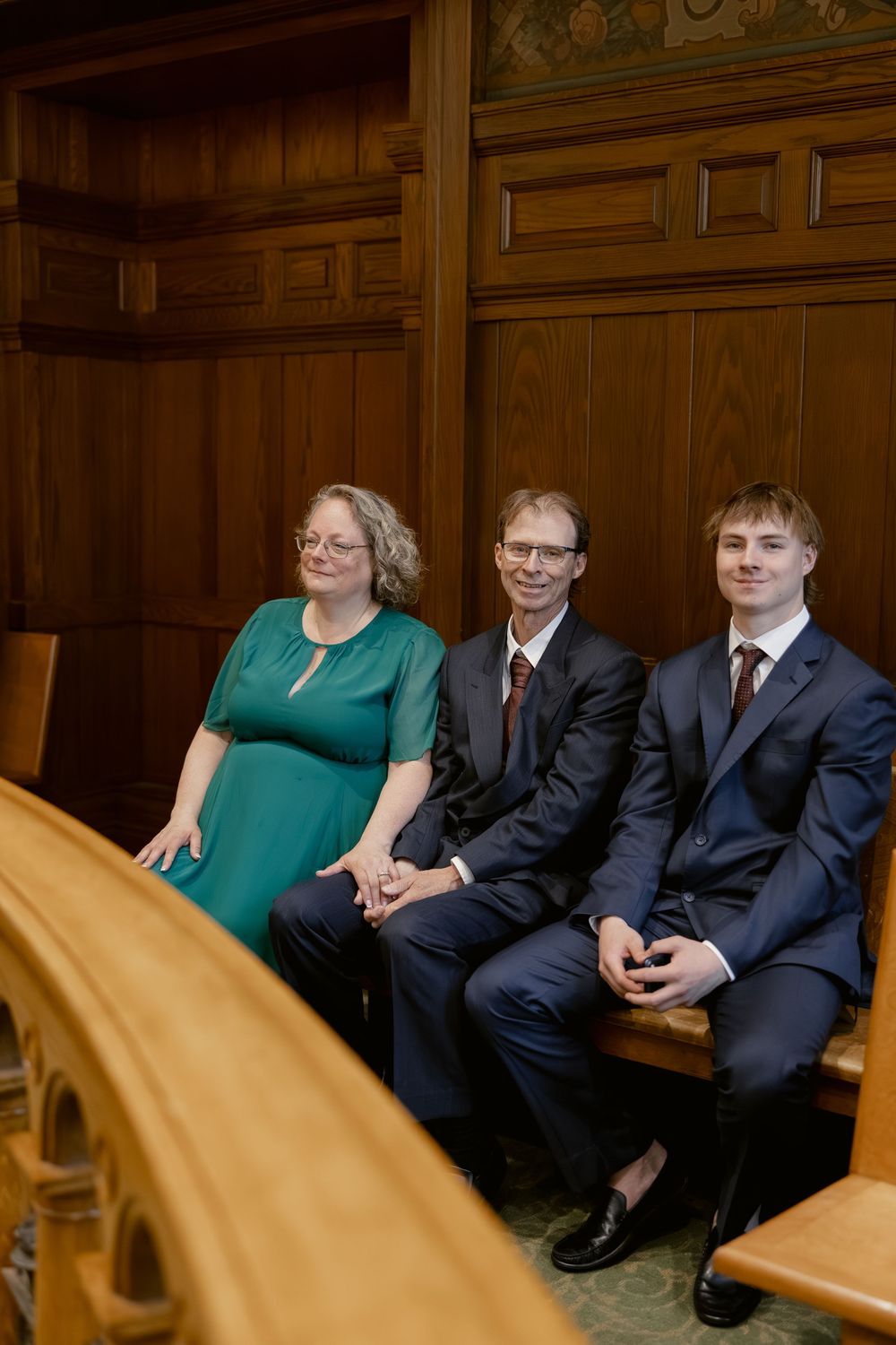 Three wedding guests, woman in teal dress and two men in suits, seated in courthouse ceremony pews.