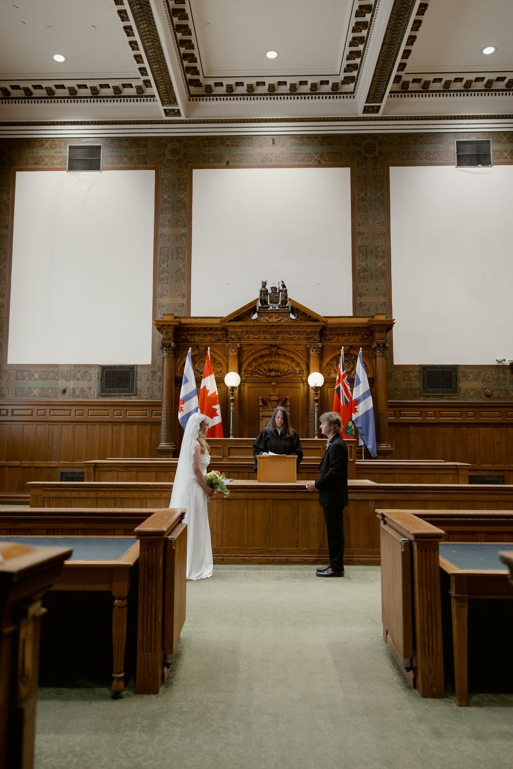 Wide-angle view of bride and groom exchanging vows before officiant in grand historic courthouse courtroom.