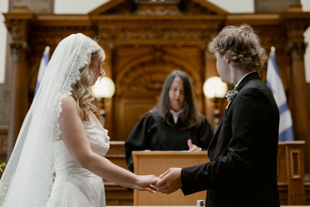 Bride and groom hold hands during wedding ceremony before officiant in elegant wood-paneled courthouse.