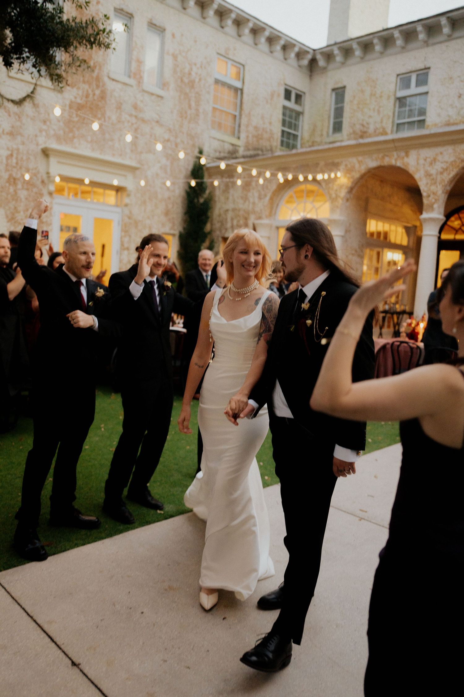 Bride in white gown and groom in tuxedo walk through cheering guests at elegant outdoor wedding venue.