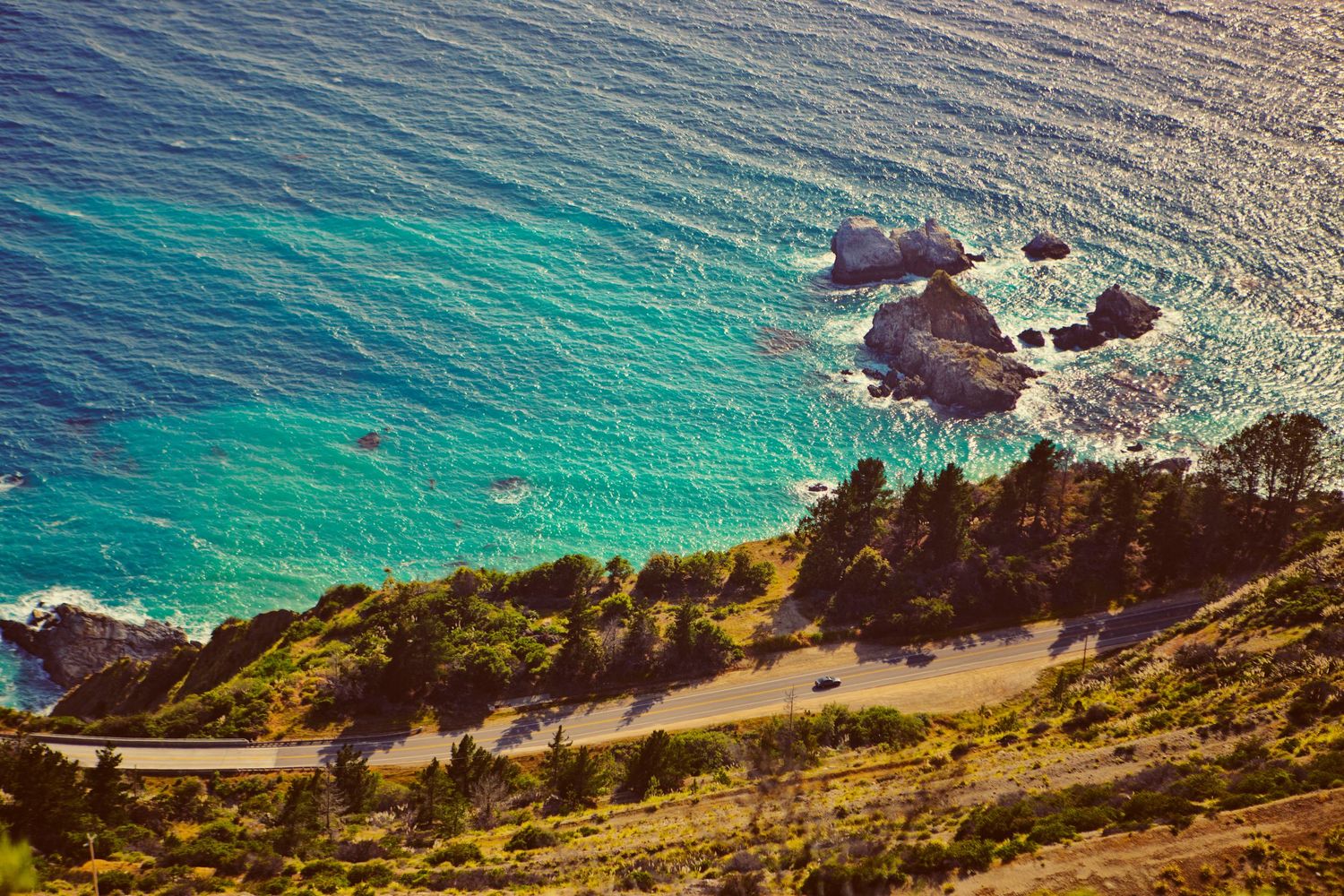 Aerial view of a stunning turquoise coastline with rocky outcrops, lush trees, and a winding coastal road in Big Sur, California.