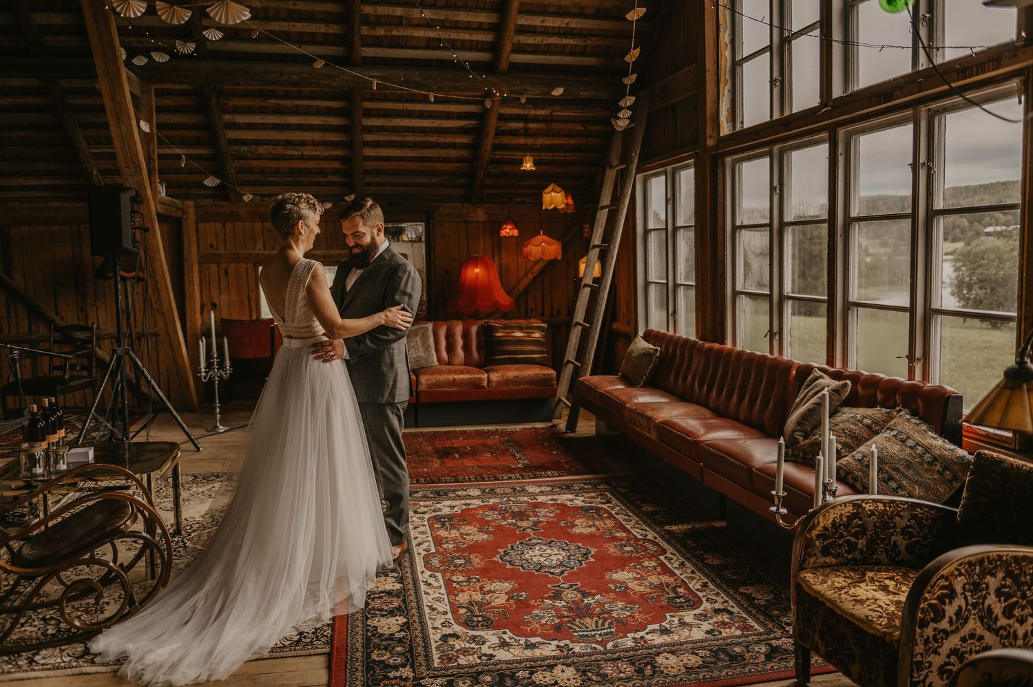 Bride and groom share a romantic moment in a rustic wooden venue with large windows and vintage decor. First look.