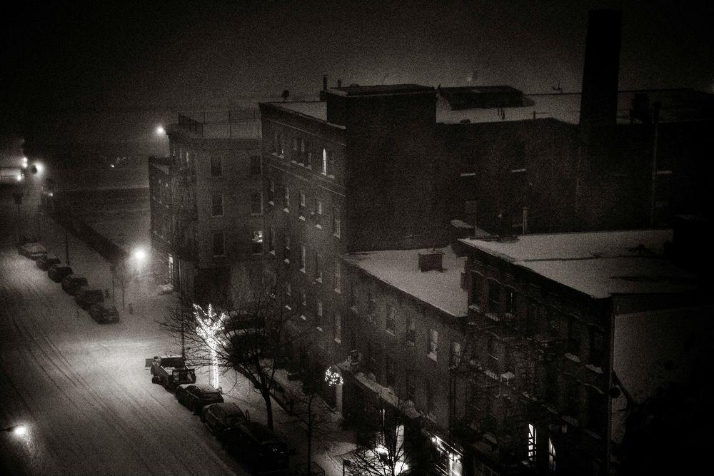Snow-covered city street at night with glowing Christmas tree lights and illuminated buildings in dark winter scene.