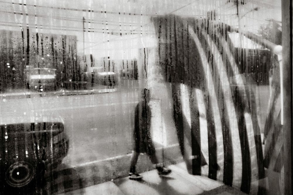 Blurry black and white photo of a person walking past a rain-soaked window with an American flag reflection.