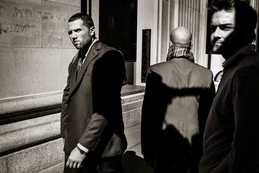 Black and white street photo of three men in suits near a building, with dramatic shadows on the wall.
