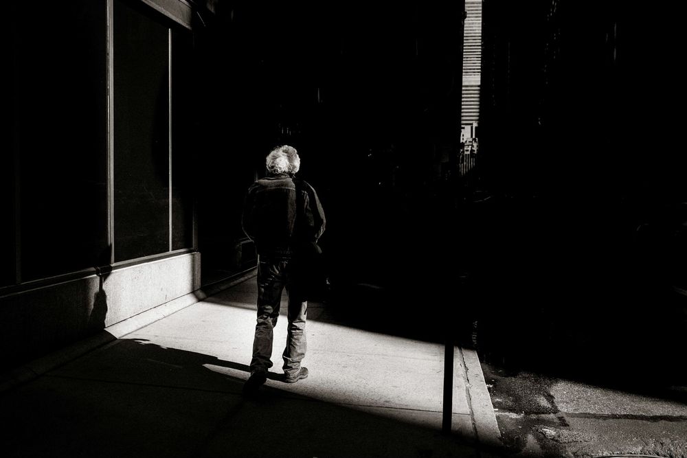 Black and white photo of a person standing in a shadowy alley with dramatic light contrast on the sidewalk.
