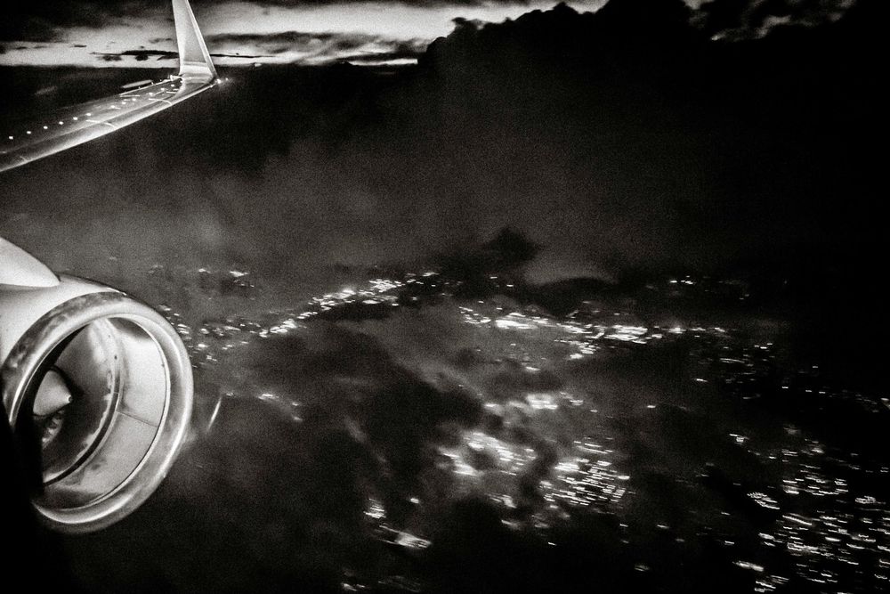 Black and white aerial view from airplane window showing engine, dramatic clouds, and city lights below at night.