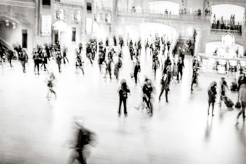 Blurred black and white photo of a busy crowd moving through a grand public hall or train station.