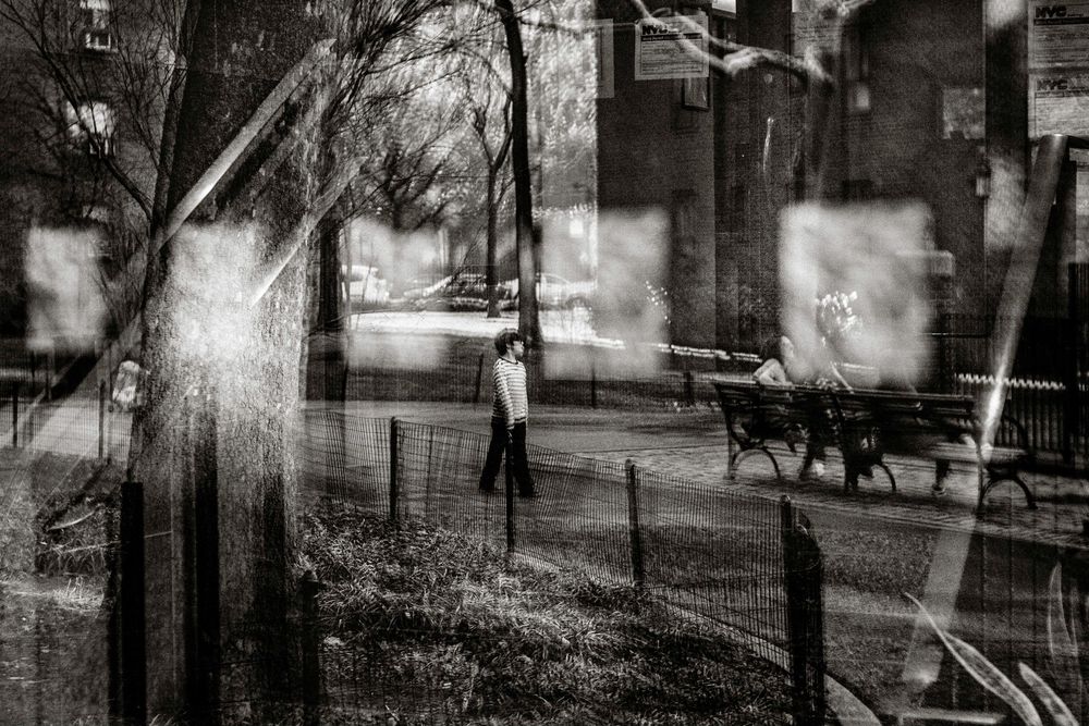 Black and white photo of a lone figure walking through an urban park with bare trees and benches in moody, atmospheric light.