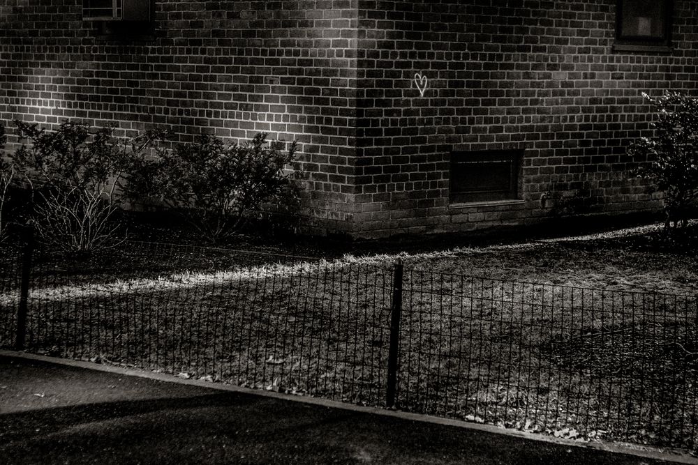 Black and white photo of a brick building with chain-link fence and shrubs in a dimly lit outdoor setting.